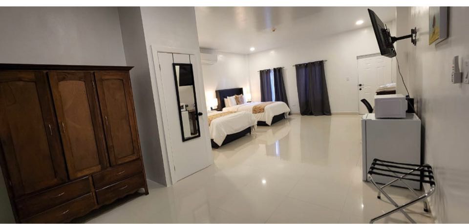 A spacious hotel room featuring two beds, a wooden wardrobe, a mini fridge, and large windows with dark curtains.