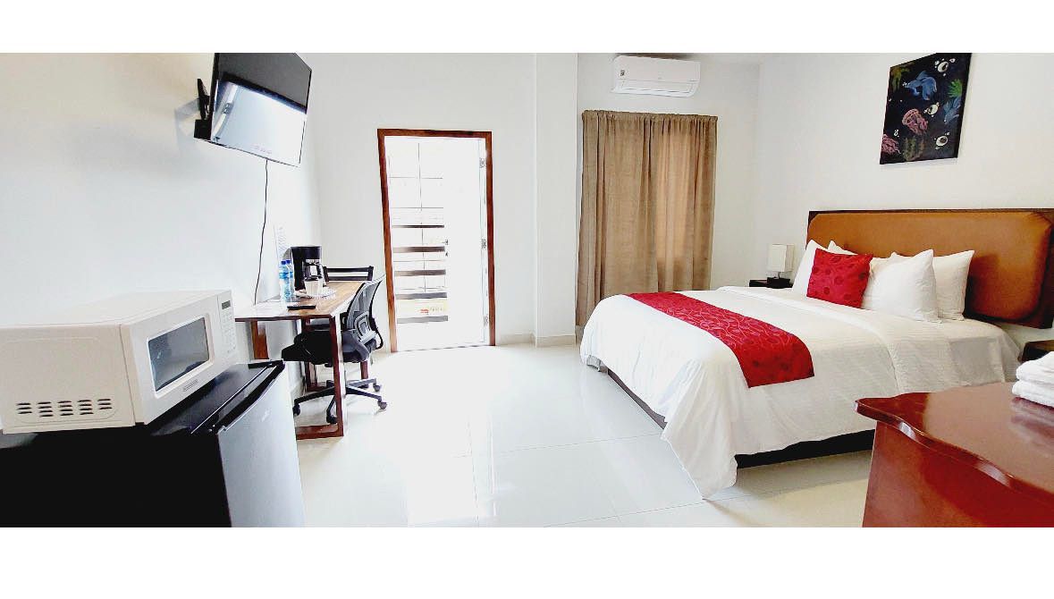 A modern hotel room featuring a bed with red accents, a desk, microwave, and TV.