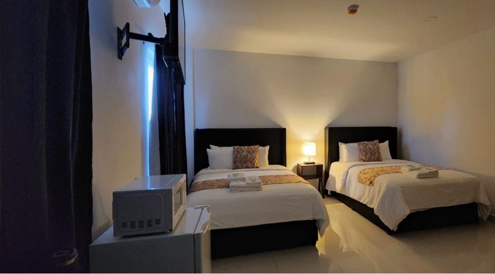 A brightly lit hotel room featuring two twin beds, a bedside lamp, and a small refrigerator.